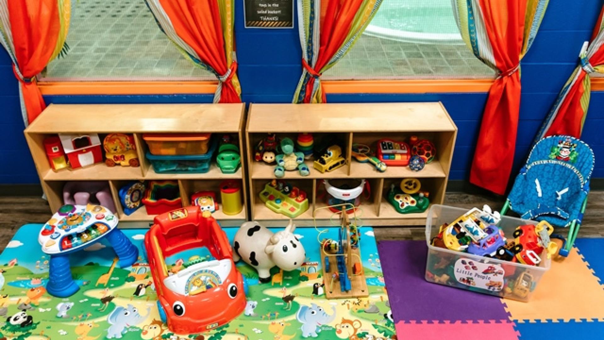 Colourful play area with toys on shelves and the floor. The floor has a foam mat and a colourful rug. There are orange drapes by the windows on the background wall.