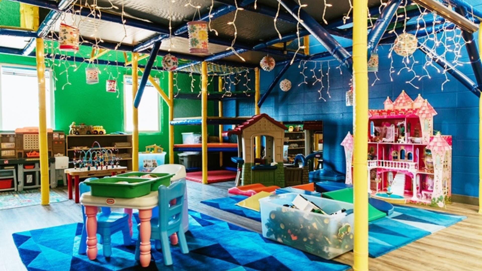 Interior of playground. There are tables, chairs, dollhouses and a bookshelf in the room. The room is decorated with hanging lights and colourful rugs.