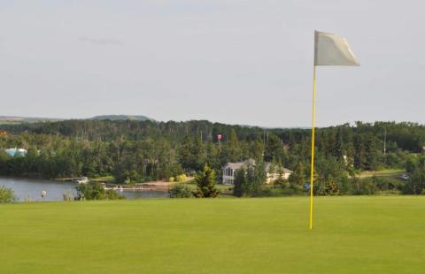 View of Hardisty Lakeview Golf Course greenway. There is a flag pin on marking the hole. In the background there are some hills and some outlines of buildings.