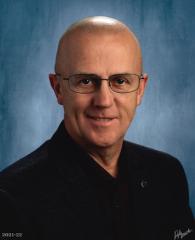 Headshot of Kevin Kinzer. There is a bald man with glasses and a black shirt in front of a blue-ish background.